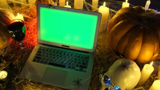 Dark scary vampire crypt, laptop with green screen against Halloween attributes, pumpkins, burning candles, glass of blood, skull glowing eyes and spiders. Halloween party, technology and social media