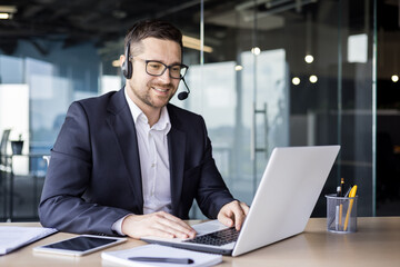 A young businessman is talking in the office on the news of a call from a laptop. He sits in the...