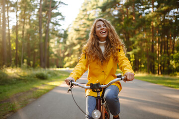 A smiling female tourist in a yellow coat enjoys the weather in the autumn park while riding a bicycle. Autumn fashion. Concept of relaxation, nature. - obrazy, fototapety, plakaty