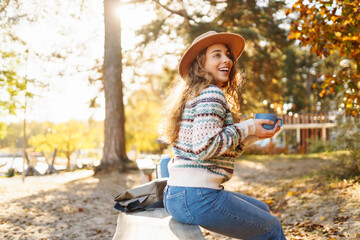 A happy woman in a stylish sweater sits on a bench in an autumn park, drinks a hot drink from a thermos and enjoys the view of the lake. Vacation concept, weekend. Lifestyle.