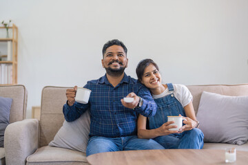 Indian couple watch tv on the couch at their living room, husband and wife spending quality time...