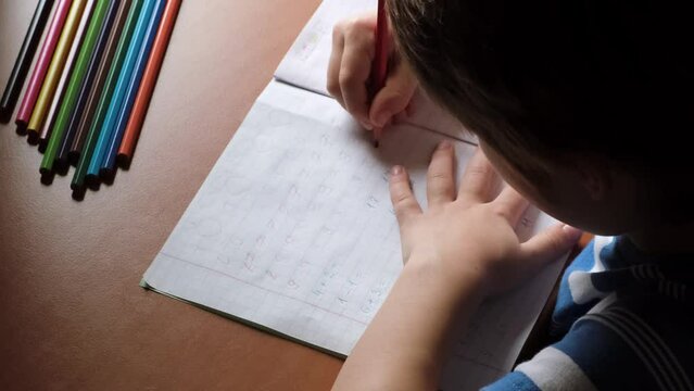 The child writes numbers in a copybook with a colored pencils. A mathematics homework for preschooler. A boy diligently learns to write. The kid is sitting at the table and doing a task. Smart person.