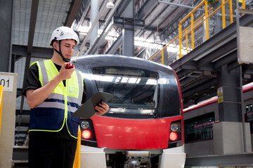 Fototapeta premium Young caucasian engineer man or worker hold clipboard for looking and checking electric train for planning maintenance and using radio for command in station, inspector check service transport.
