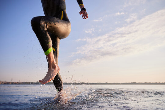 Cropped image of male body, leg in wetsuit running out of river after training in early morning. Triathlete workout. Concept of professional sport, triathlon preparation, competition, athleticism