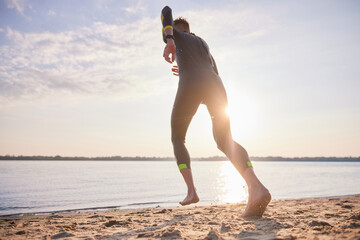 Sportive man in wetsuit running on beach into water for early morning training. Swimming,...