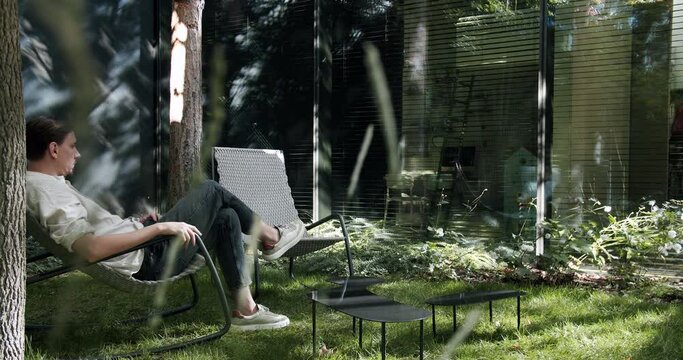 Young man sitting and resting in lounge chair outdoors near modern cottage house at sunny day. Male relax in armchair in backyard of vacation home. Weekend on nature concept