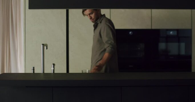 Young man in casual clothes turn on faucet and filling glass with cold water to drink at kitchen with modern minimalist interior.
