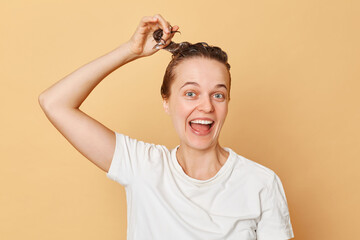 Hair care and hygiene. Pampering routine. Overjoyed excited woman wearing white T-shirt washes hair standing with shampoo foam on her head isolated over beige background