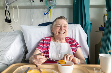 Happy older woman patient laughing in her NHS hospital bed, London, England, UK