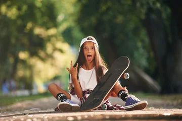 Fotobehang Front view, showing gestures. Happy little girl with skateboard outdoors © standret
