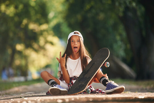 Front view, showing gestures. Happy little girl with skateboard outdoors