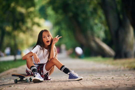 Having fun, showing hand gesture. Happy little girl with skateboard outdoors