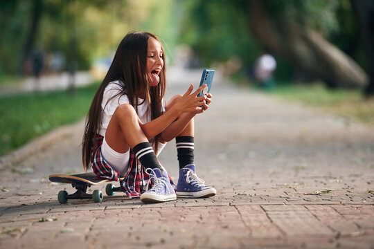 Showing tongue, making selfie. Happy little girl with skateboard outdoors