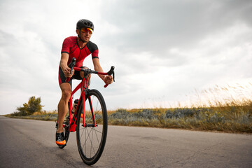 Competitive and motivated man, triathlete riding bicycle on road outdoors, training for marathon. Concept of professional sport, triathlon preparation, competition, athleticism - obrazy, fototapety, plakaty
