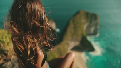 Womans hair flutters in the wind slow motion. Back view of tourist girl sitting on the rock with ocean view. Travel, tourism, holiday. Indonesia, Nusa Penida Island, Kelingking Secret Point Beach