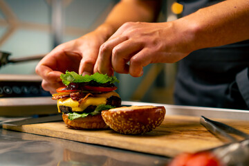 A close-up of a chef preparing a burger with beef patty with vegetables and cheese in the professional kitchen of an Italian restaurant - obrazy, fototapety, plakaty