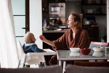 Mother wearing bathrope spoon feeding her infant baby boy child sitting in high chair at the dining table in kitchen at home in the morning. - 649311649