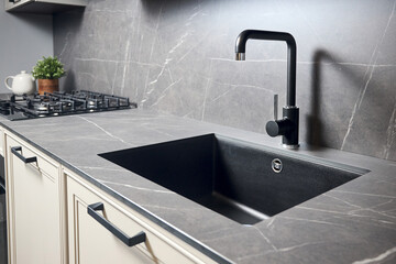 Compact undermount sink. Kitchen sink area with black square matte sink tap in contemporary style. Matte black and stoneware kitchen design. Black ceramic sink with gas hob and oven in background.