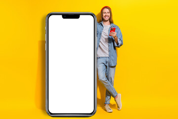 Full size photo of handsome cheerful man wear jacket jeans trousers lean on touchscreen smartphone isolated on yellow color background