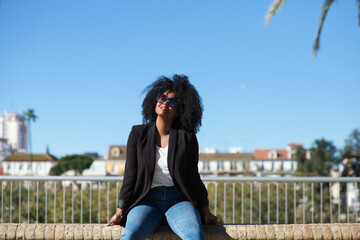 Fototapeta premium Young beautiful black woman with afro hair sitting on a bench wearing jeans and black jacket enjoying her holidays in spain. Travel and holiday concept.