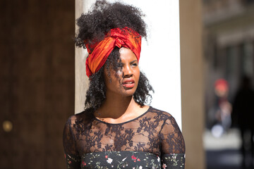 Portrait of a young, beautiful black woman with afro hair and black dress with flowers, wearing a red scarf in her hair. The woman is sad and serious and leaning on a marble column. - Powered by Adobe