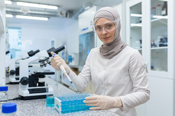 Portrait of a young Muslim woman in hijab working in a laboratory, sitting at a desk with samples...