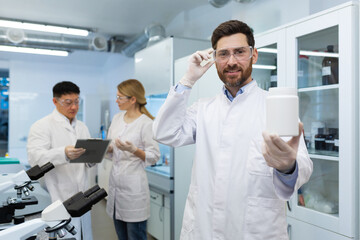 Portrait of a smiling young man pharmacist standing in a laboratory in a uniform and holding a plastic jar with material in his hands. Shows the result to the camera. Colleagues are discussing behind.