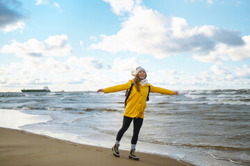 A female traveler in a yellow coat and backpack walks and has fun on the ocean shore, enjoying the scenery. The concept of freedom, travel, recreation. Lifestyle.