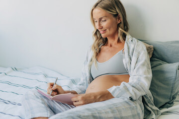 A pregnant woman writes in a notepad while sitting on the bed.