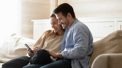 Happy man and woman using phone together, sitting on couch, watching video in social network or...