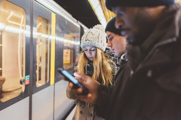 three young adult friends waiting metro and checking smartphone