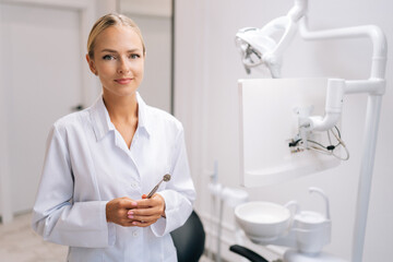 Portrait of attractive female dental doctor holding dental equipment for work and looking at camera in dentistry clinic with modern white interior. Concept of stomatology, dentistry and orthodontics.