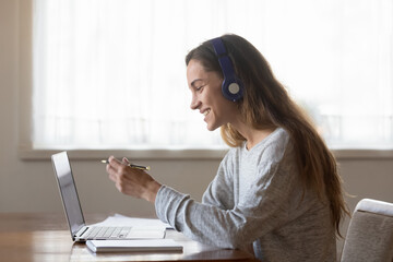Smiling woman wearing headphones looking at laptop screen, holding pen, writing notes during...
