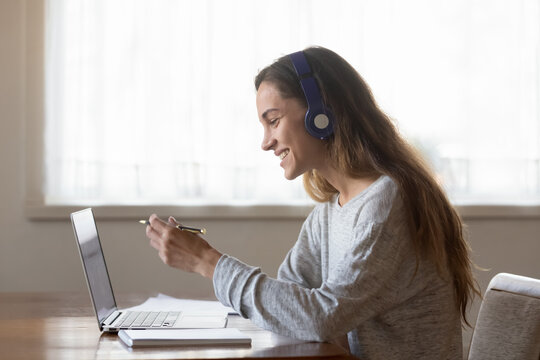 Smiling woman wearing headphones looking at laptop screen, holding pen, writing notes during webinar, happy female student sitting at desk, learning language online, teacher holding online lecture