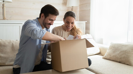 Happy young couple unpacking awaited parcel at home together, curious man and woman looking into...