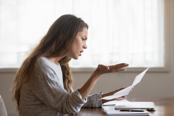 Angry annoyed woman reading unexpected bad news in letter, holding paper, sitting at work desk,...