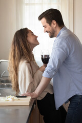 Happy young couple drinking red wine, standing near table in modern kitchen, clinking glasses, looking at each other with love, family celebrating wedding anniversary or romantic date