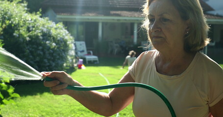 Older woman gardening and water plants with water hose outside in sunlight