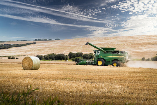Rocky View County Alberta Canada, September 18 2023: John Deere combine harvesting a wheat field signals a full hopper of grain as it passes a round straw bale with Jet trails across a prairie sky.
