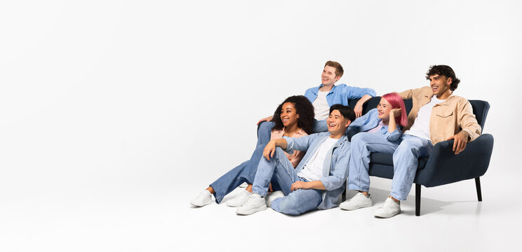 Excited diverse teen friends watching tv, sitting on sofa and floor on white background, looking at copy space, panorama - Powered by Adobe