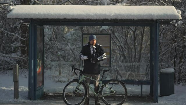 Frozen male cyclist waiting for bus at halt stop with bicycle in winter in snowy weather. Velocipede broke down and man waits long time for transport at the bus stop in winter after a snow storm. 