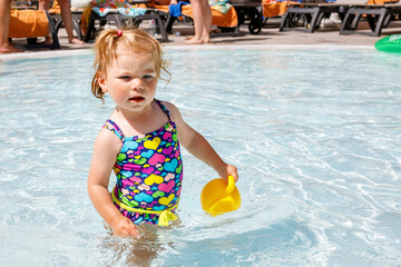 Naklejka premium Cute happy little toddler girl in the pool and having fun on family vacations in a hotel resort. Healthy child playing in water. Baby in colorful fashion swimsuit.