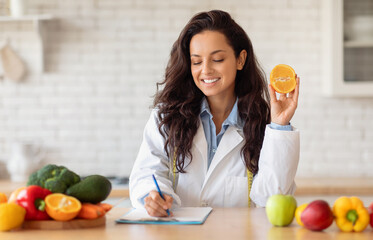 Cheerful european lady doctor nutritionist in white coat writing diet plan, recipe with organic...