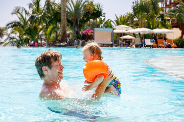 Cute happy little toddler girl and father in the pool and having fun on family vacations in a hotel resort. Healthy child and man playing in water. Baby daughter in colorful fashion swimsuit.