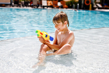 Happy little kid boy jumping in the pool and having fun on family vacations in a hotel resort. Healthy child playing in water with a water gun. Laughing running child splashing