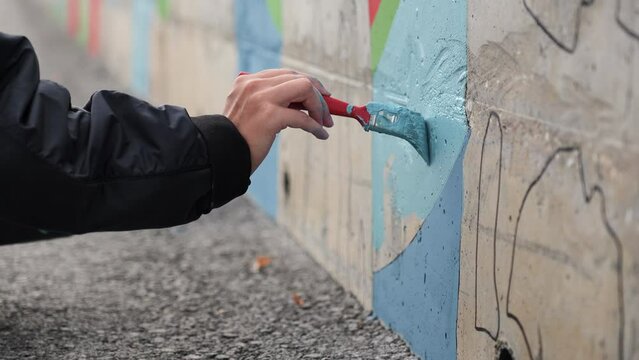 Male teenager hand painting a wall in blue color with a paint brush. Clip. Talented student creating street art object.