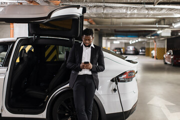 Fototapeta na wymiar Relaxed african american adult in business suit sending message on phone while waiting for electric car to be recharged. Efficient manager chatting with colleague via gadget in bottom-level parking.