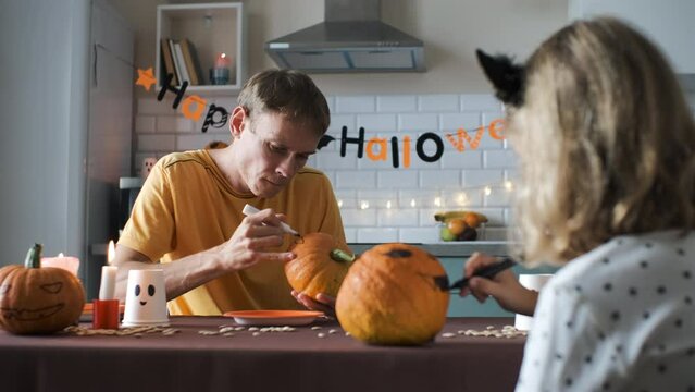 Father with his little daughter painting scary faces on pumpkins while sitting at table in the kitchen. Halloween preparation concept