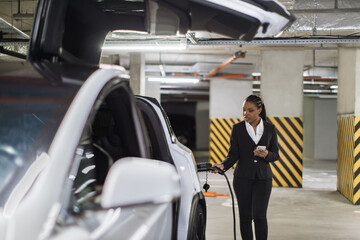 Focus on attractive lady with cell phone standing near electric automobile with charge cable in...