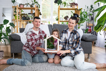 Traditional family with child are enjoying new home, sitting on the sofa and holding the roof. Mortgage, insurance and protection, buying and moving into your own home, green house with potted plant